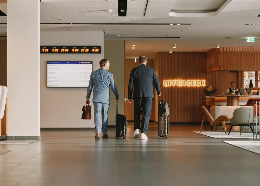 Two guests with suitcases are walking next to the reception in the lobby. The entrance to the UPPER DECK bar is visible.